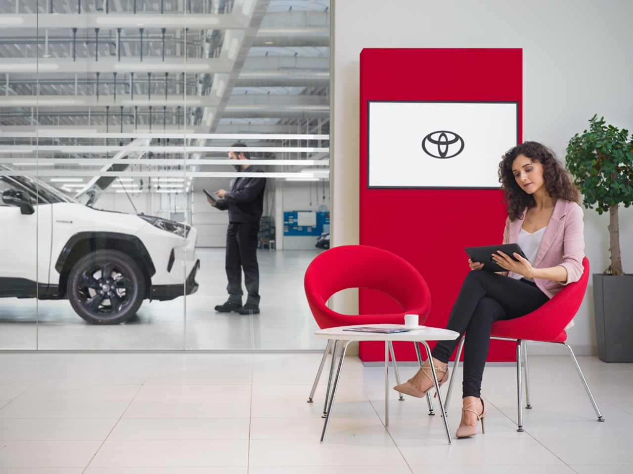 A women sits in a Toyota Service waiting area. She looks at a tablet, with a Toyota logo displayed on a tv screen behind her.